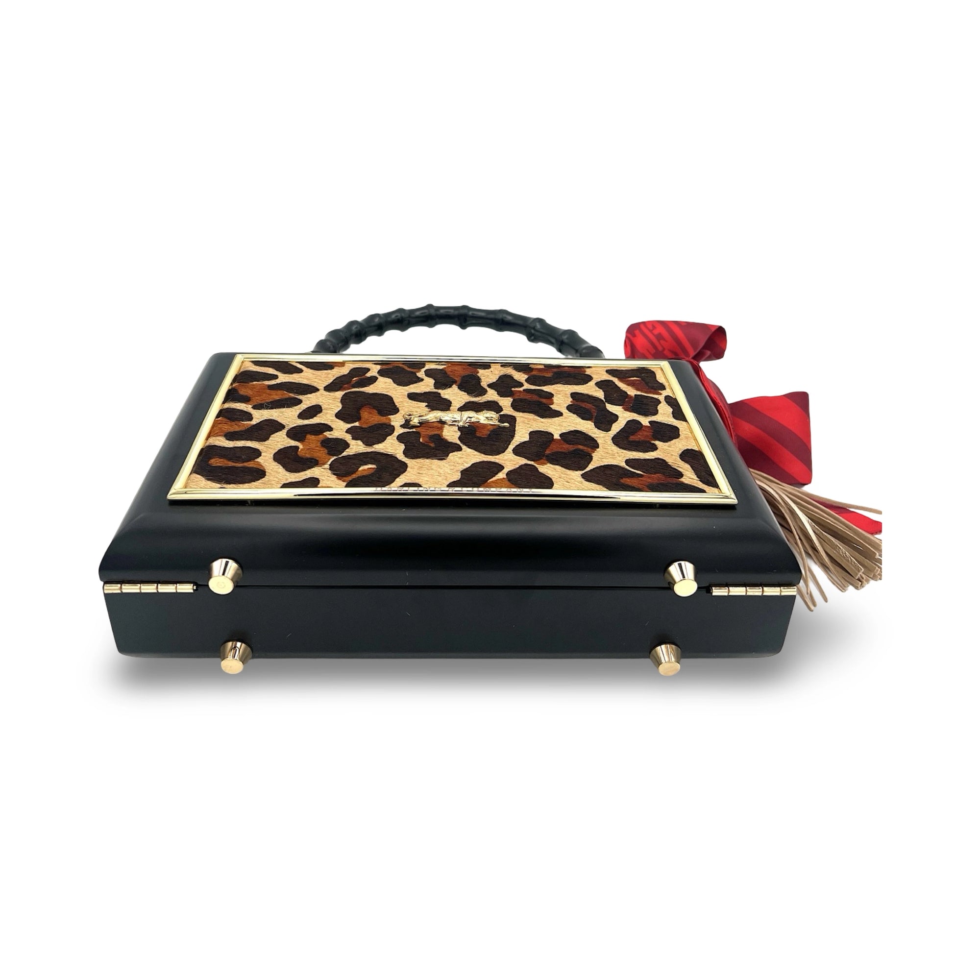 The Leopard Supreme Bag by Faire features a rectangular leopard print cowhide design, gold accents, a textured black bamboo handle, and a red bow with a gold tassel on the side, displayed against a white background.