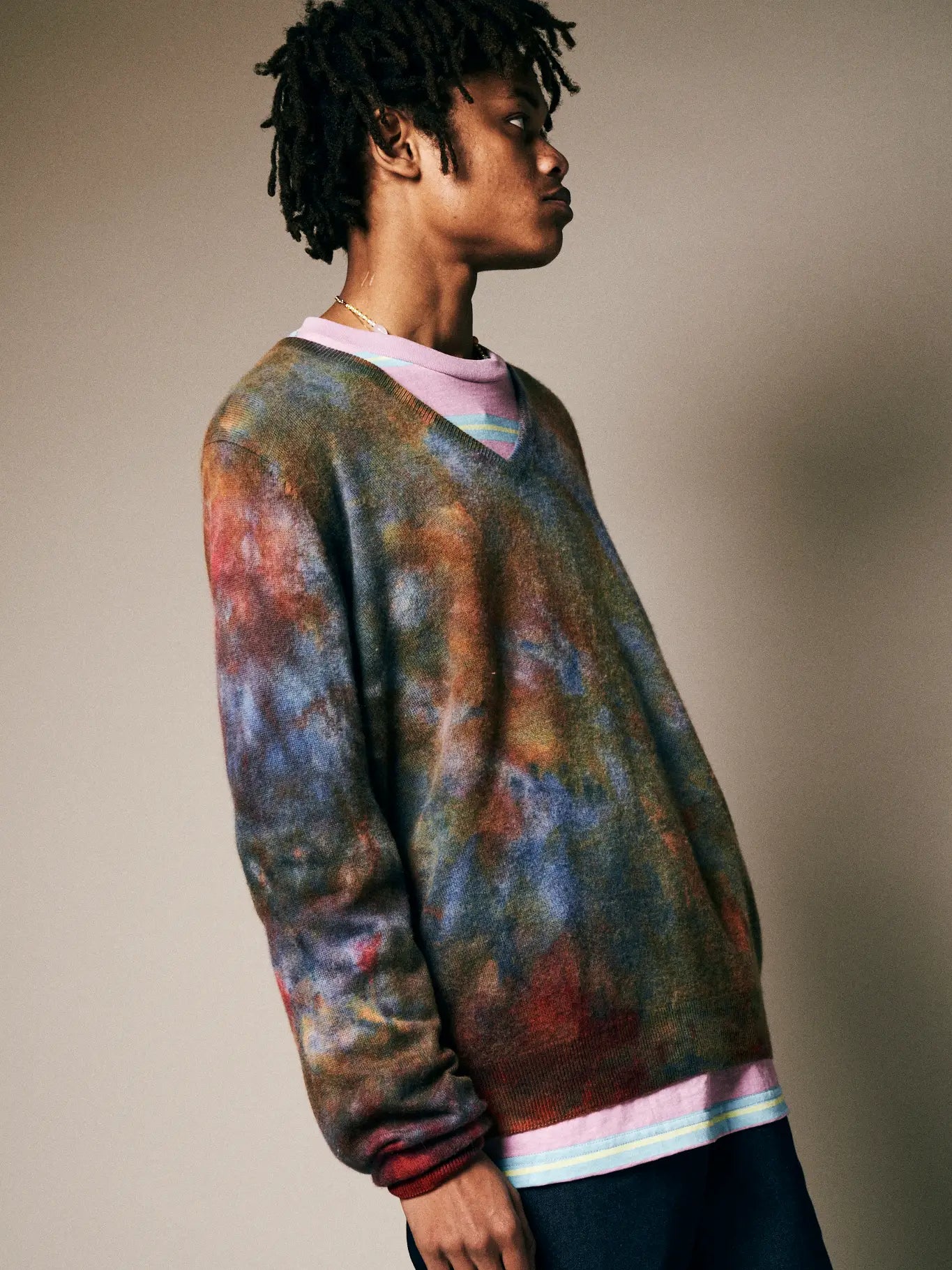 A person with short dreadlocks stands sideways against a neutral background, wearing the Riverside Tool & Dye Cashmere V-neck over a pink shirt.