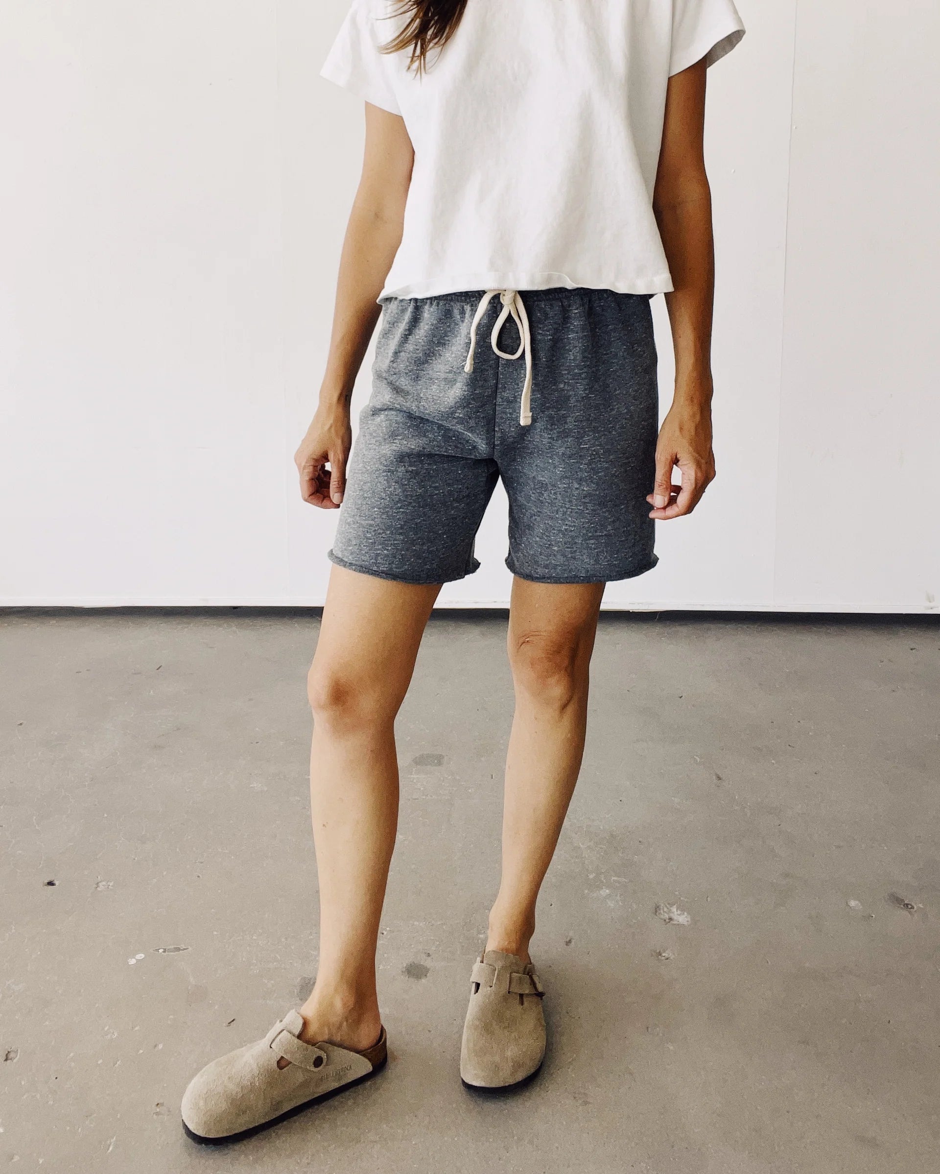 A person stands indoors on a concrete floor, wearing Hey Gang’s The Sweat Shorts in vintage-inspired cotton French terry, a white T-shirt, and beige slip-on shoes. The photo is cropped at the shoulders with a plain white background.