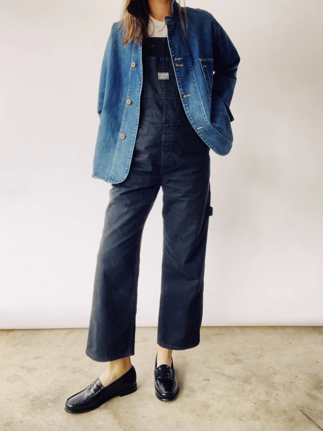 A person is wearing The Barn Coat by Hey Gang, paired with black overalls and loafers, standing against a plain background. The person's head is not visible in the image.