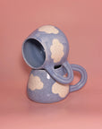Two Cute Mugs by Faire, handmade with blue glaze and white cloud patterns, are stacked on a pink background—one resting sideways atop the other.