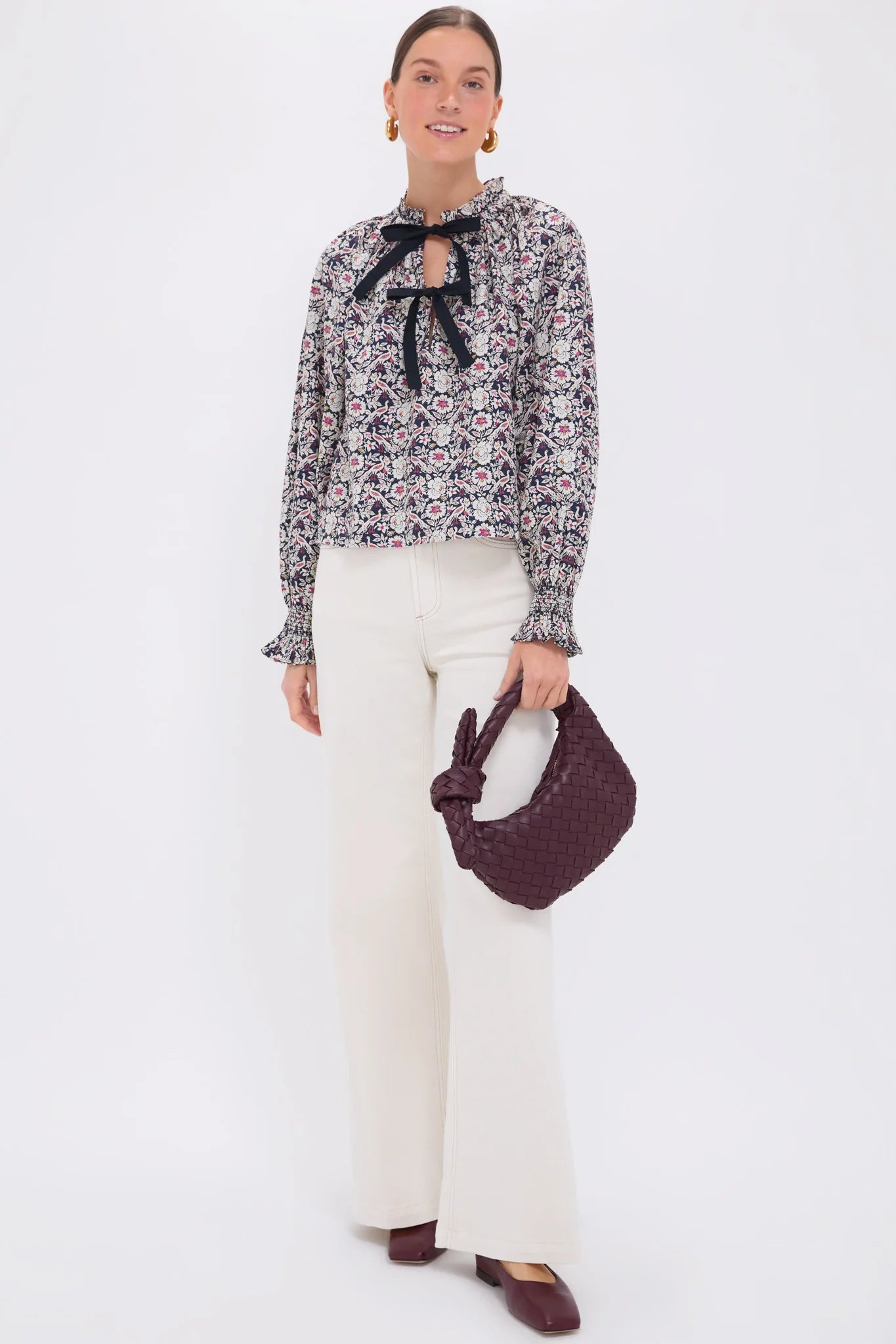 Against a plain backdrop, a person models the Rhode Aiden Top, featuring a ruffle collar and black self-tie bow. They pair it with cream wide-leg pants, burgundy shoes, and matching woven bag. Gold hoop earrings adorn them as their hair is neatly pulled back.