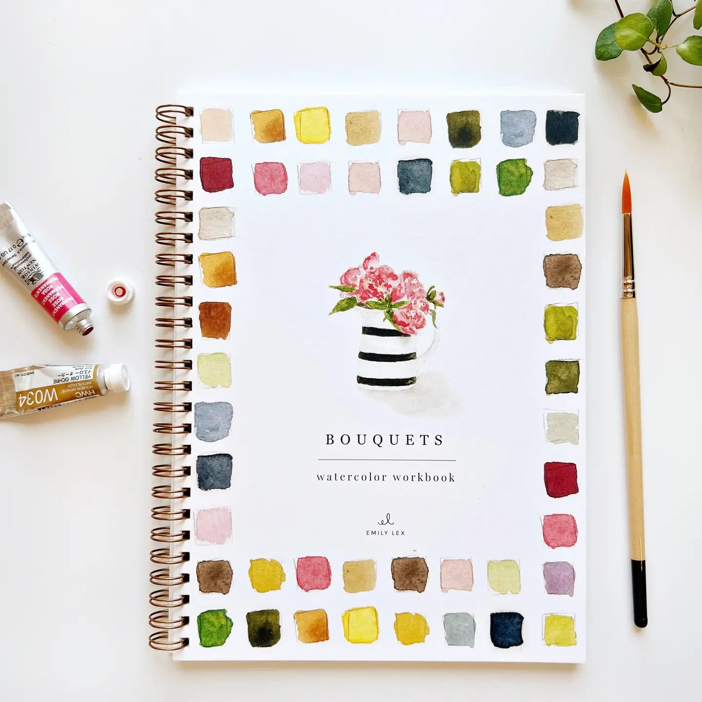 Faire's Watercolor Workbook is a beginner-friendly, spiral-bound guide featuring a flower vase illustration with colorful paint swatches around the border. Paint tubes, a brush, and a small plant accent the white surface nearby.