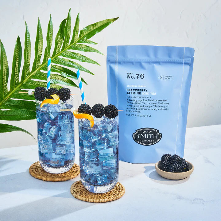 Two glasses of Faire Blackberry Jasmine Iced Tea sit on woven coasters. Next to them are a blue bag of tea sachets and a small bowl of blackberries, with a green leaf visible in the background.