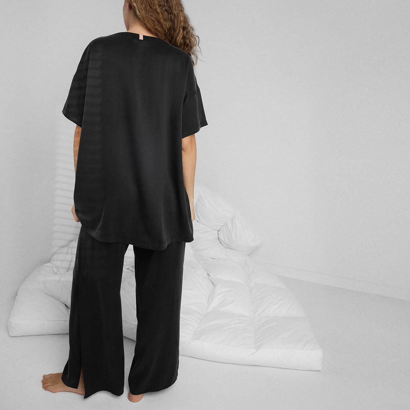 A person with long curly hair stands barefoot on a white floor, facing away and wearing Faire’s Lunya Washable Silk Pant with side slits. A rumpled white comforter is on the floor in the background.