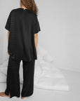 A person with long curly hair stands barefoot on a white floor, facing away and wearing Faire’s Lunya Washable Silk Pant with side slits. A rumpled white comforter is on the floor in the background.