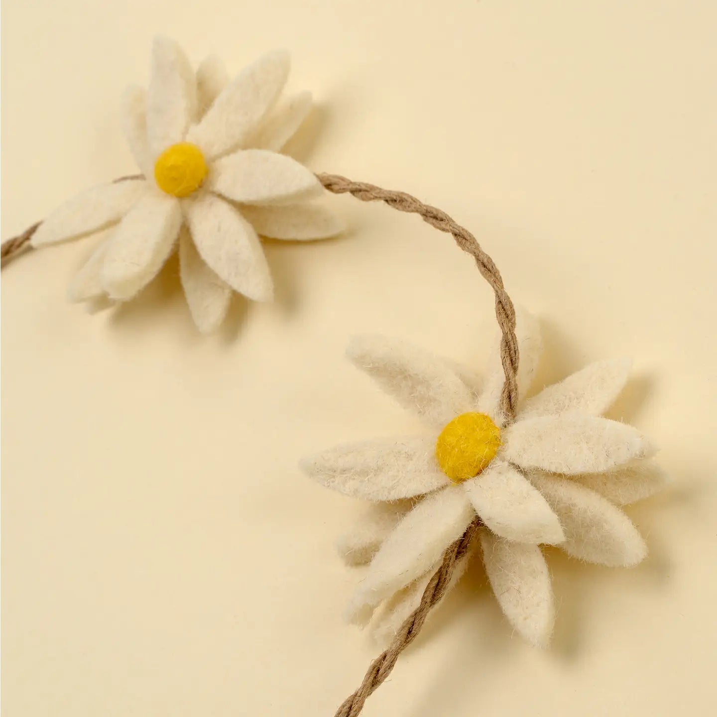 A close-up of Indaba's Daisy Flower Garland shows two handmade felt daisies with yellow centers and white petals on a twisted brown string, ideal for sustainable home decor, set against a light cream background.