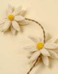 A close-up of Indaba's Daisy Flower Garland shows two handmade felt daisies with yellow centers and white petals on a twisted brown string, ideal for sustainable home decor, set against a light cream background.