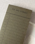 The Faire To Do Today Planner is a green 50-sheet notepad with hourly slots from 9 AM to 5 PM. It sits on a light textured surface alongside a pen, postage stamps, and an open envelope.