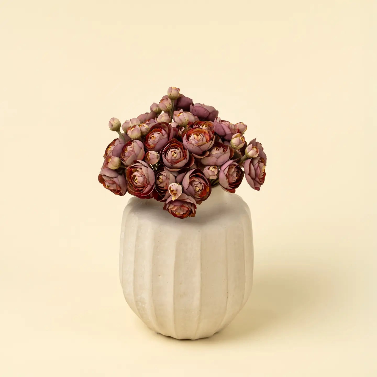The Indaba Ranunculus Bouquet features mauve and pink blooms in a white, ridged ceramic vase, perfect for displaying decorative vases against a plain, light beige background.