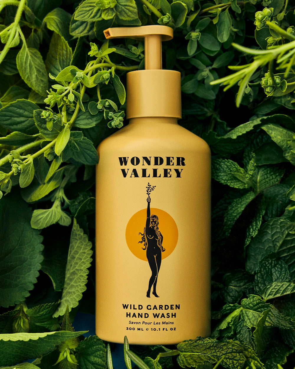 A yellow bottle of Faire Wild Garden Hand Wash rests among green leaves, featuring bold black text and an illustration of a woman holding a branch, evoking its herbal scent and deep cleansing benefits.