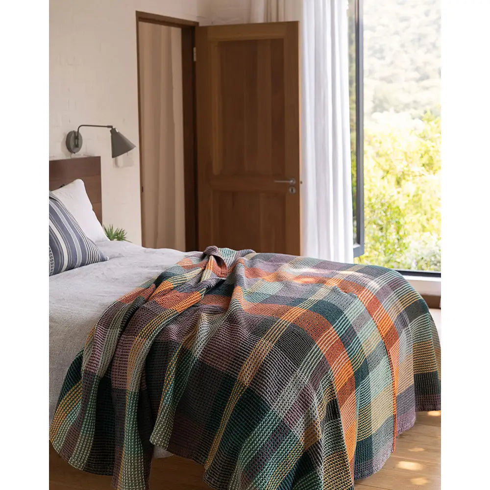 A cozy bedroom features a wooden bed with white bedding, topped with a colorful Faire Vrou-Vrou Blanket. An open door and large window fill the space with natural light and views of greenery outside.