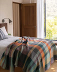 A cozy bedroom features a wooden bed with white bedding, topped with a colorful Faire Vrou-Vrou Blanket. An open door and large window fill the space with natural light and views of greenery outside.