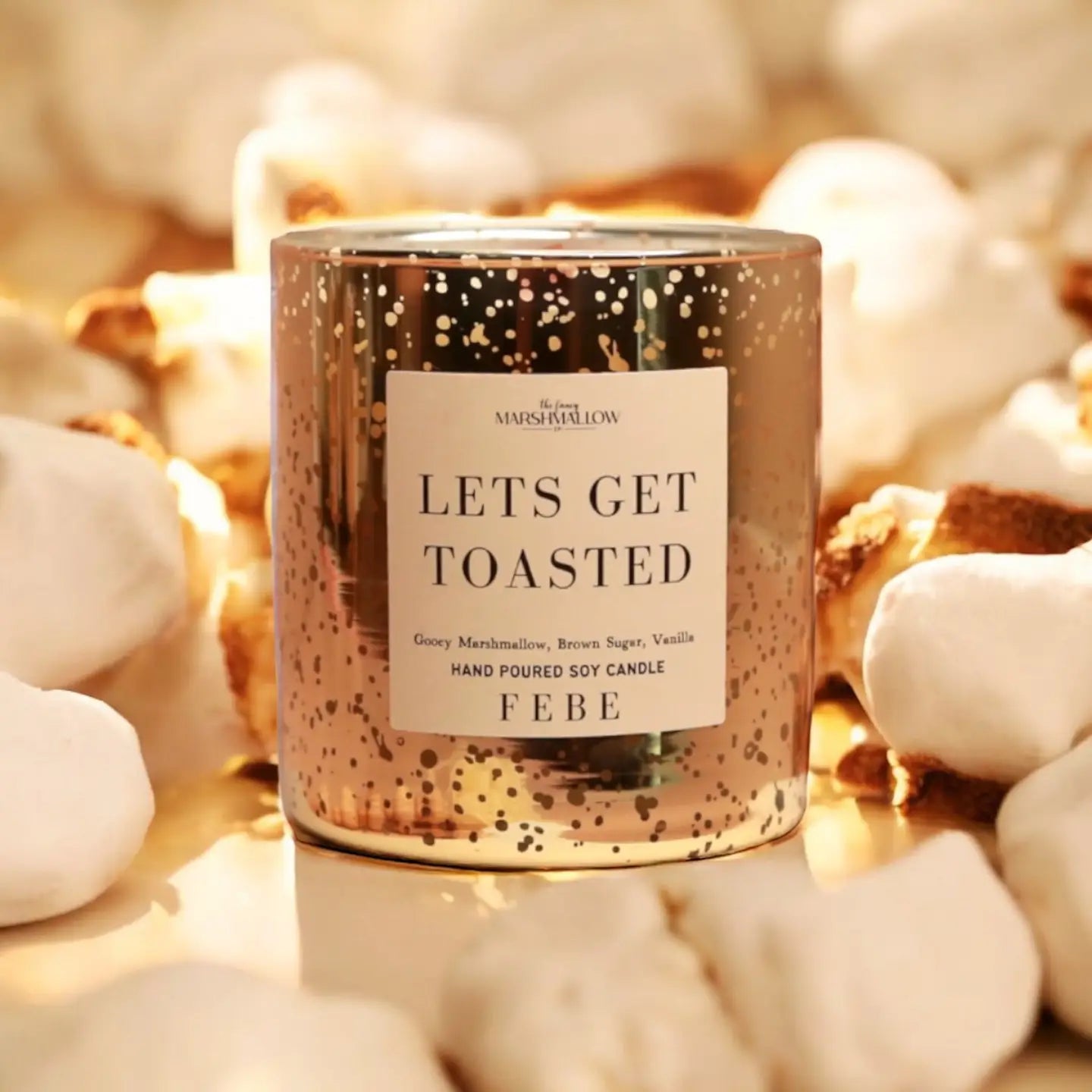 A gold candle from Faire labeled "Let's Get Toasted" is surrounded by toasted marshmallows and showcases a speckled design. It emits scents of gooey marshmallow, brown sugar, and vanilla. This Smoke Show creation is hand-poured by FEBE Candles.