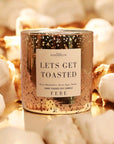 A gold candle from Faire labeled "Let's Get Toasted" is surrounded by toasted marshmallows and showcases a speckled design. It emits scents of gooey marshmallow, brown sugar, and vanilla. This Smoke Show creation is hand-poured by FEBE Candles.