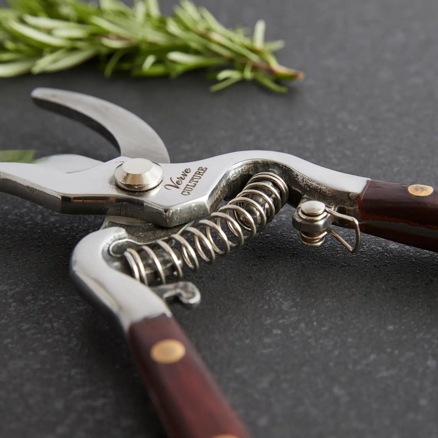 Close-up of Faire Thai Kitchen Garden Shears with wooden handles and sharp stainless steel blades, lying open on a dark surface. A sprig of rosemary rests nearby.
