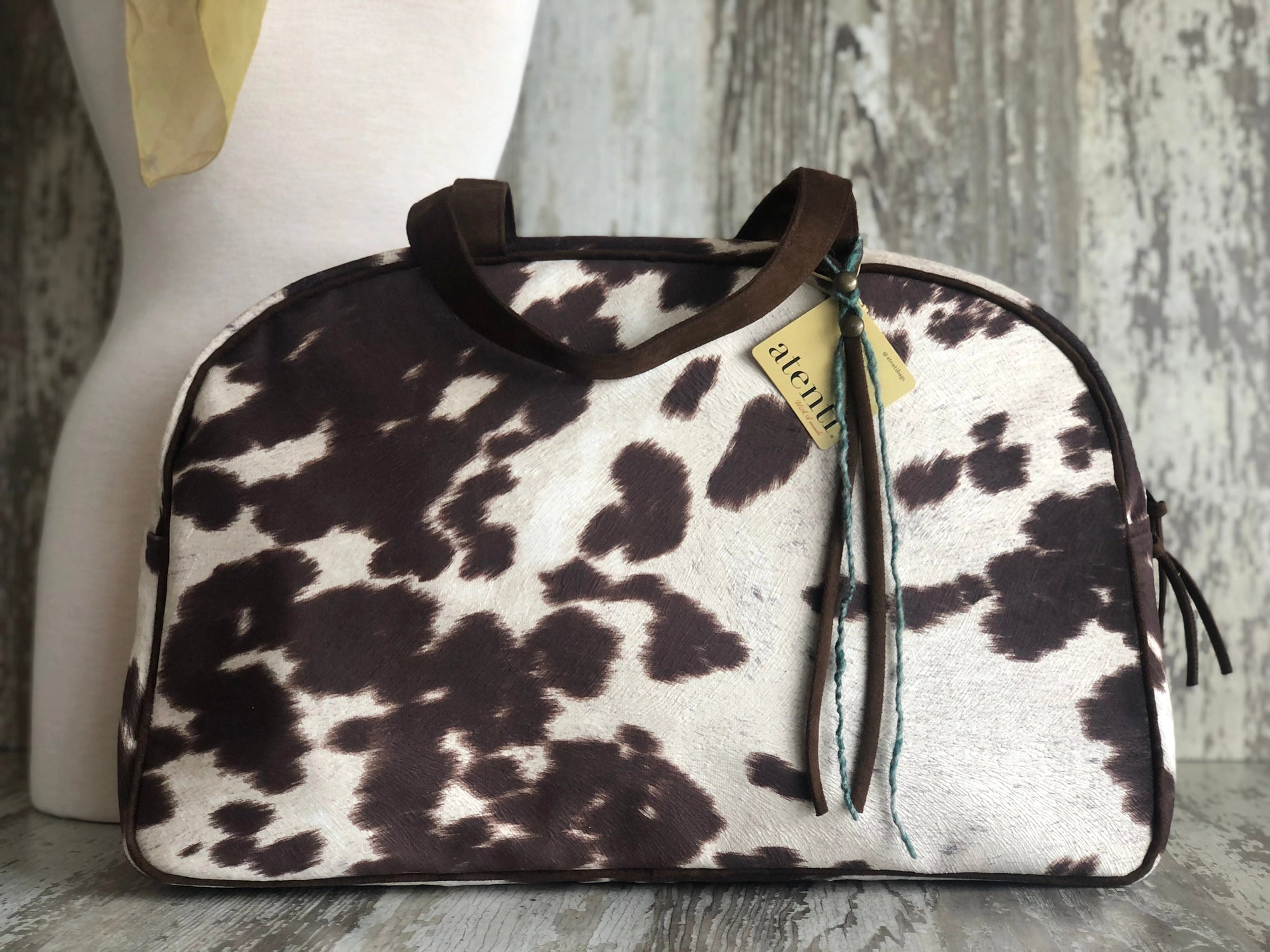 The Oakley Overnighter by Faire is a handmade USA handbag featuring a faux cowhide brown and white pattern, dark brown handles, a yellow "alenvi" tag, and ribbon accents, displayed against a rustic wooden background.