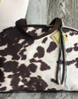 The Oakley Overnighter by Faire is a handmade USA handbag featuring a faux cowhide brown and white pattern, dark brown handles, a yellow "alenvi" tag, and ribbon accents, displayed against a rustic wooden background.