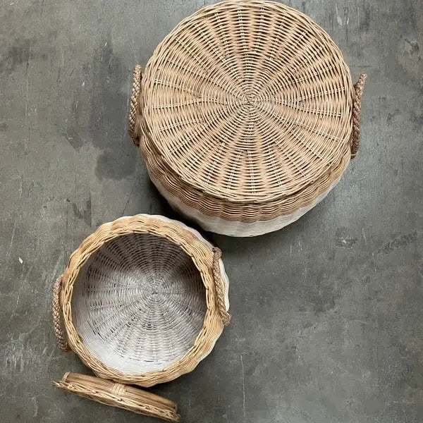 Two Faire Lidded Round Baskets with straw side handles rest on a gray concrete surface; one is closed, and the other reveals its woven white and natural interior. Both baskets combine style and function for versatile storage.
