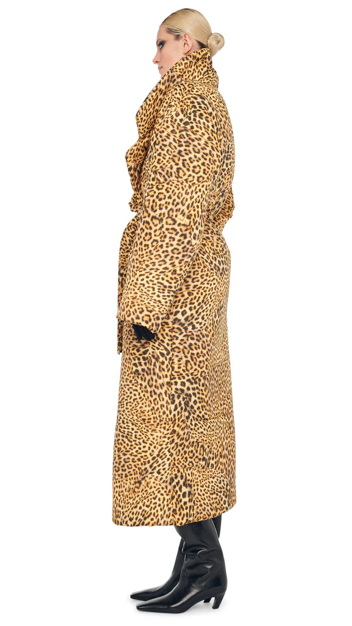 A person stands in profile view, confidently wearing the Classic Sleeping Bag Coat by Norma Kamali, featuring a long leopard print design. The coat is belted at the waist and paired with black boots, while their hair is pulled back elegantly.