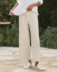 Standing outdoors amidst lush greenery and a stone patio, a person wears a white long-sleeve shirt, Frank & Eileen's Wexford The Trouser in cream—a high-rise, wide-leg linen pant paired with white sneakers. Their face remains unseen.