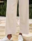 A person stands outdoors on a stone path, wearing Frank & Eileen's Wexford The Trouser. The cream-colored high-rise wide-leg linen pants beautifully complement the lush green shrubs in the background and are paired with white sneakers.