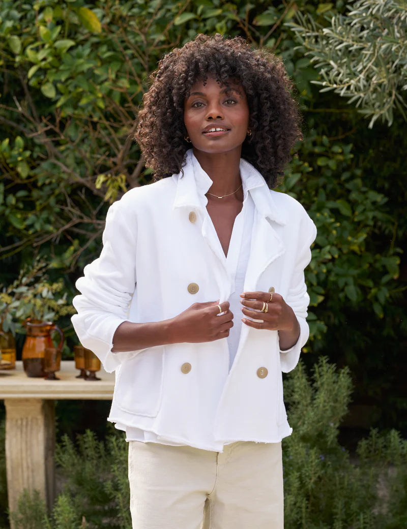 A woman with curly hair wears a Frank & Eileen MINI BELFAST Crop Peacoat. Its fit enhances her style as she stands outside, smiling with hands in pockets adorned with horn buttons. The backdrop includes lush greenery and a table featuring amber glass bottles.