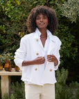 A woman with curly hair wears a Frank & Eileen MINI BELFAST Crop Peacoat. Its fit enhances her style as she stands outside, smiling with hands in pockets adorned with horn buttons. The backdrop includes lush greenery and a table featuring amber glass bottles.