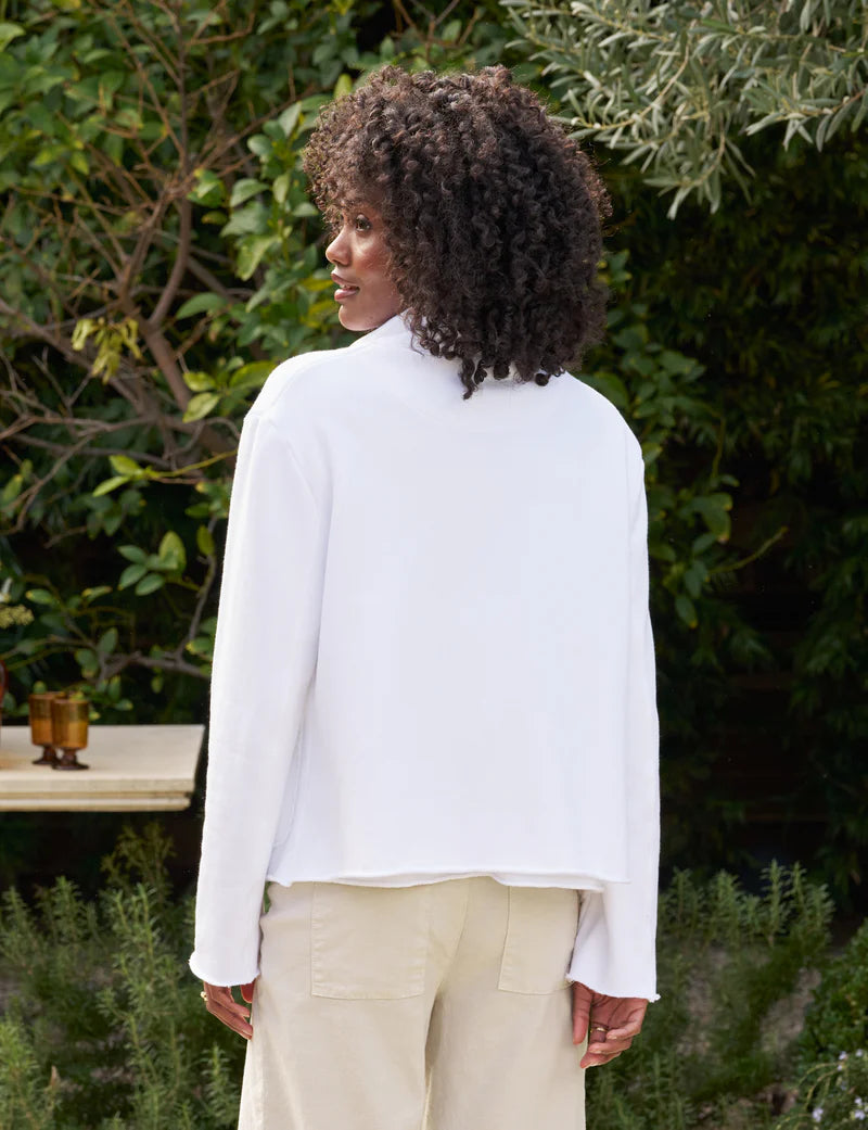 A person with curly hair stands outdoors, facing away. They're wearing a cropped fit white MINI BELFAST Crop Peacoat by Frank & Eileen and beige pants, surrounded by lush greenery and a wooden table with a candle holder in the background.