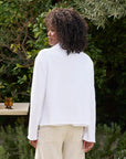 A person with curly hair stands outdoors, facing away. They're wearing a cropped fit white MINI BELFAST Crop Peacoat by Frank & Eileen and beige pants, surrounded by lush greenery and a wooden table with a candle holder in the background.