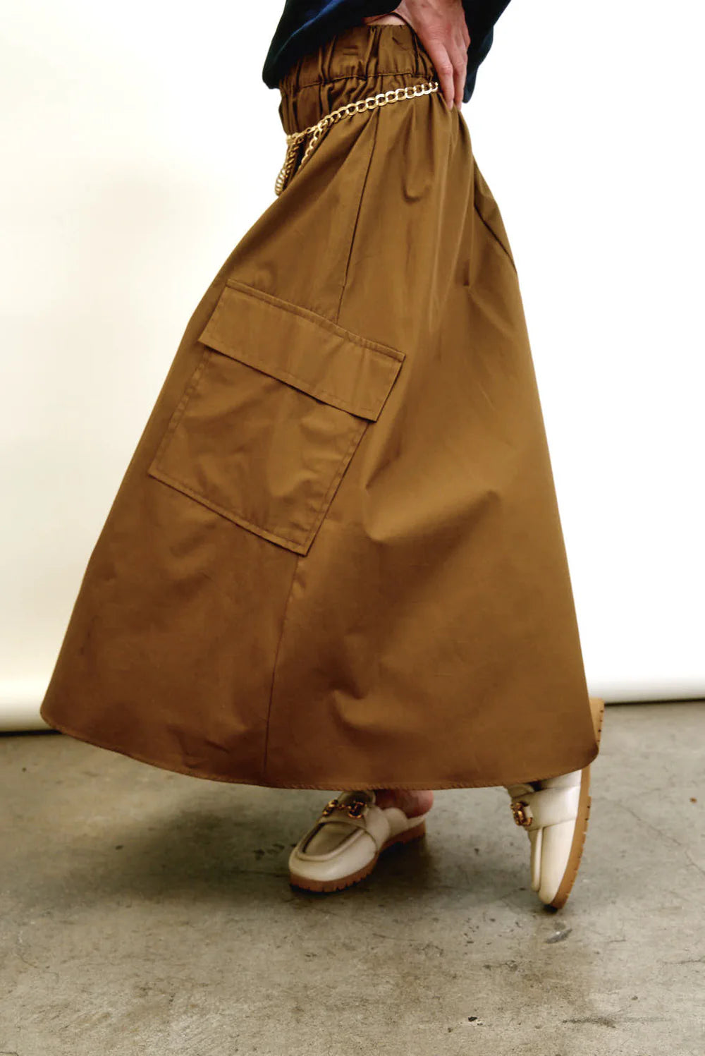Model wears the Aquarius Cocktail Le Cargo Skirt— a long brown skirt with oversized pockets—styled with a gold chain belt and beige loafers featuring gold accents, standing on a concrete floor against a plain light background.