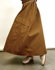 Model wears the Aquarius Cocktail Le Cargo Skirt— a long brown skirt with oversized pockets—styled with a gold chain belt and beige loafers featuring gold accents, standing on a concrete floor against a plain light background.