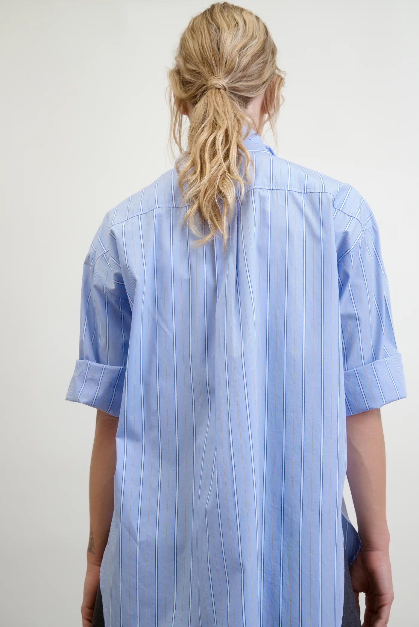 A person with long, wavy blonde hair is seen from the back, wearing a Dear Polly Shirting Bow Blouse in light blue with classic mens shirting stripes and rolled-up sleeves. The Japanese Cotton Shirting from Aquarius Cocktail adds a crisp elegance to their look. The background is plain and neutral.