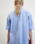 A person with long, wavy blonde hair is seen from the back, wearing a Dear Polly Shirting Bow Blouse in light blue with classic mens shirting stripes and rolled-up sleeves. The Japanese Cotton Shirting from Aquarius Cocktail adds a crisp elegance to their look. The background is plain and neutral.