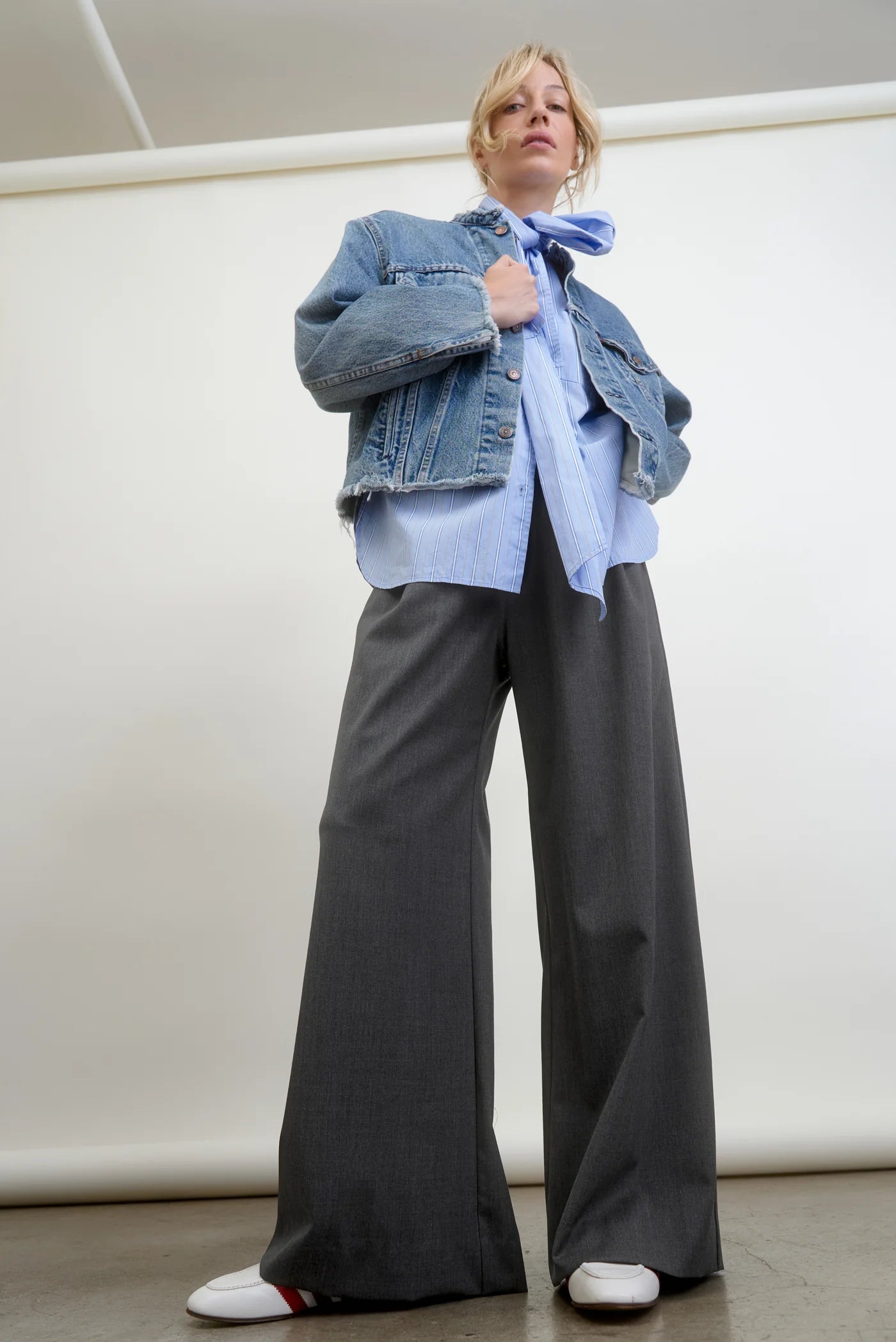 A person stands against a plain backdrop, wearing a denim jacket over a blue shirt, paired with the Connor Wide Leg Pant in gray wool blend from Aquarius Cocktail, and white sneakers. The overall fashion style is casual and modern, highlighting small-batch made pieces for a unique flair.