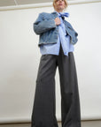 A person stands against a plain backdrop, wearing a denim jacket over a blue shirt, paired with the Connor Wide Leg Pant in gray wool blend from Aquarius Cocktail, and white sneakers. The overall fashion style is casual and modern, highlighting small-batch made pieces for a unique flair.