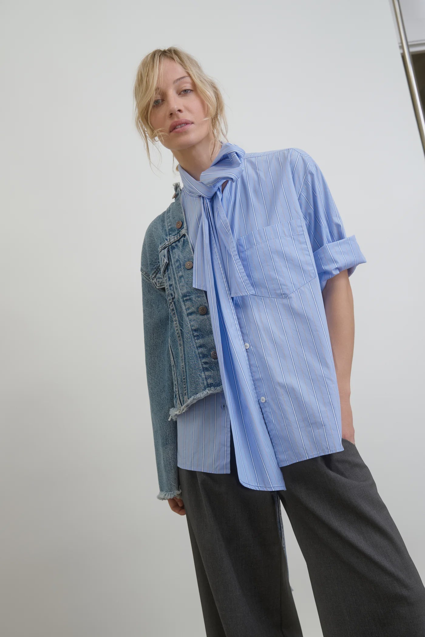 A person with blond hair is wearing the Dear Polly Shirting Bow Blouse in light blue from Aquarius Cocktail, styled with a scarf tied around the neck and layered with a denim jacket. The sleeves are rolled up, and the person is paired with dark gray trousers. The minimalist background creates a perfect face-framing effect.