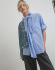 A person with blond hair is wearing the Dear Polly Shirting Bow Blouse in light blue from Aquarius Cocktail, styled with a scarf tied around the neck and layered with a denim jacket. The sleeves are rolled up, and the person is paired with dark gray trousers. The minimalist background creates a perfect face-framing effect.