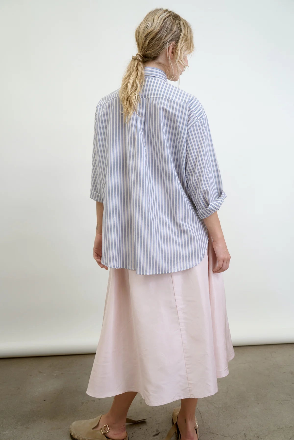 Facing away, a person with long, wavy hair tied in a low ponytail sports the Aquarius Cocktail's blue and white Dear Polly Shirting Bow Blouse made from Japanese cotton shirting, paired with a light pink midi skirt and tan buckle shoes. The background is plain and light-colored.