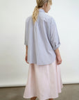 Facing away, a person with long, wavy hair tied in a low ponytail sports the Aquarius Cocktail's blue and white Dear Polly Shirting Bow Blouse made from Japanese cotton shirting, paired with a light pink midi skirt and tan buckle shoes. The background is plain and light-colored.