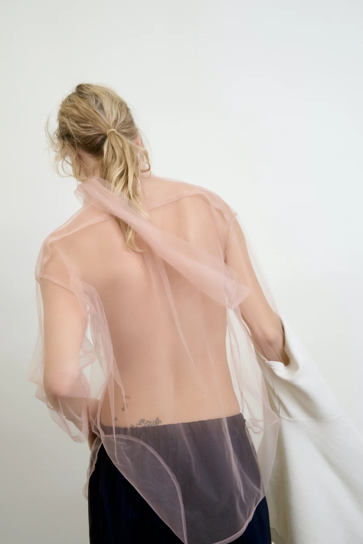 A person with long, blonde hair stands facing a light-colored wall, wearing the sheer pink Dear Polly Bow Blouse by Aquarius Cocktail. They hold a white garment over one arm and have a tattoo on their lower back.