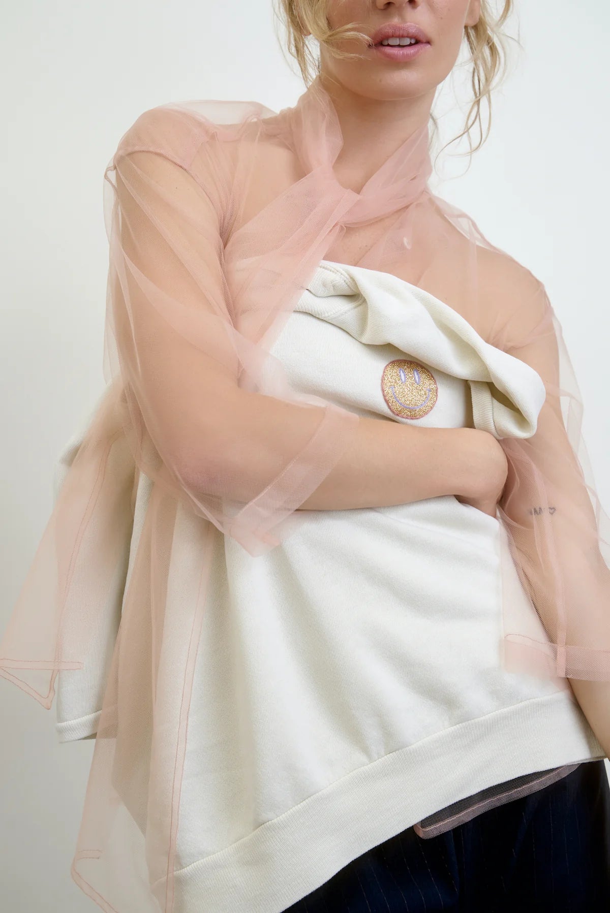 In a neutral setting, an individual is wearing the Aquarius Cocktail "Dear Polly Bow Blouse," a sheer peach-colored mesh tulle top with an oversized fit over a white garment with a pin, highlighting its delicate texture and casual style.