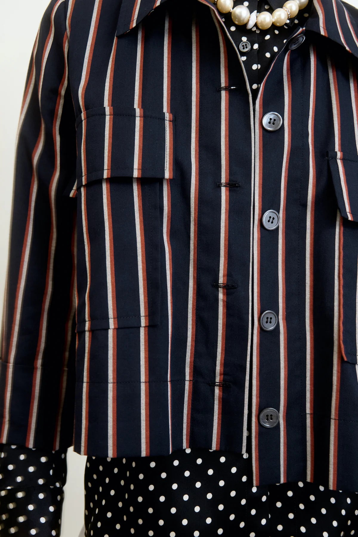 Close-up of someone wearing the Aquarius Cocktail "Yes Sir" cropped jacket in navy blue with red and white stripes over a black polka dot shirt. The jacket offers buttons, pocket flaps, and a relaxed fit, accessorized with a pearl necklace for a stylish look.