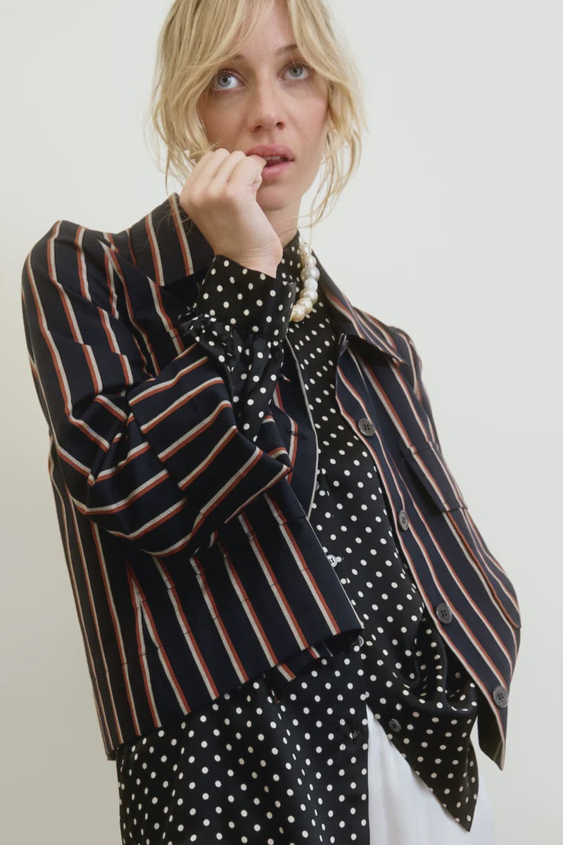 A person with blond hair wears the Yes Sir Cropped Jacket by Aquarius Cocktail, featuring stripes over a polka dot blouse. They gaze upwards with a hand on their chin, while a pearl necklace adorns their neck. The outfit's relaxed fit complements the plain, light background beautifully.