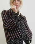 A person with blond hair wears the Yes Sir Cropped Jacket by Aquarius Cocktail, featuring stripes over a polka dot blouse. They gaze upwards with a hand on their chin, while a pearl necklace adorns their neck. The outfit's relaxed fit complements the plain, light background beautifully.