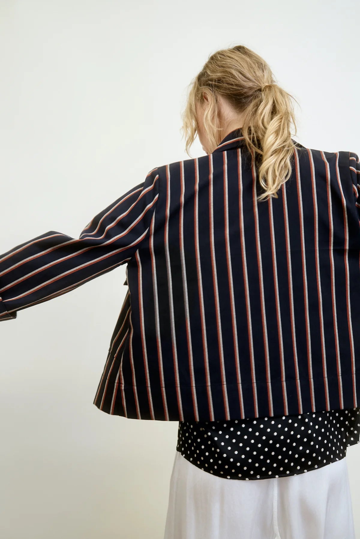 A person with blond hair faces away from the camera, wearing the "Yes Sir Cropped Jacket" by Aquarius Cocktail over a black and white polka dot shirt. They pair this with white pants, all set against a plain background.