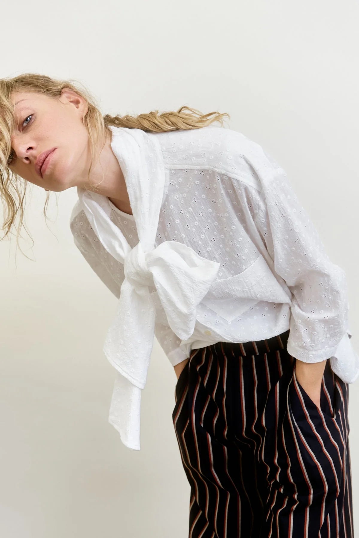 A person in the Dear Polly Shirting Bow Blouse by Aquarius Cocktail, made from small batch cotton poplin, leans forward with hands in pockets. They wear black pants with red and beige stripes, have loosely styled blonde hair, and stand against a neutral background.