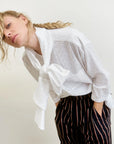 A person in the Dear Polly Shirting Bow Blouse by Aquarius Cocktail, made from small batch cotton poplin, leans forward with hands in pockets. They wear black pants with red and beige stripes, have loosely styled blonde hair, and stand against a neutral background.