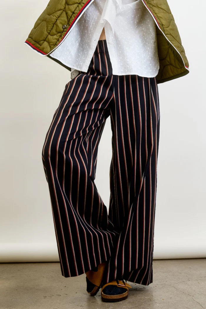 A person in Connor Wide Leg Pant by Aquarius Cocktail, featuring navy blue wool blend with red and white stripes. They wear a white shirt and green quilted jacket, standing with crossed legs to highlight black shoes with brown soles.