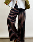 A person in Connor Wide Leg Pant by Aquarius Cocktail, featuring navy blue wool blend with red and white stripes. They wear a white shirt and green quilted jacket, standing with crossed legs to highlight black shoes with brown soles.